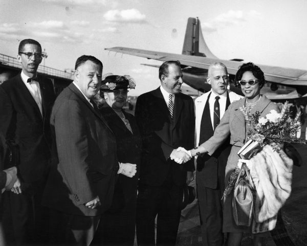 Daisy Bates as part of a welcoming party at an airport. From left to right are: Edward L. Cooper, executive secretary of the Boston NAACP, Kivie Kaplan, National Executive Board, Melnea Cass of the Boston Fighting Fund for Freedom, John D. Brown, the official greeter for the City of Boston, Herbert E. Tucker, Jr., President of Boston Branch, and Daisy Bates, who is holding flowers and a fur wrap.