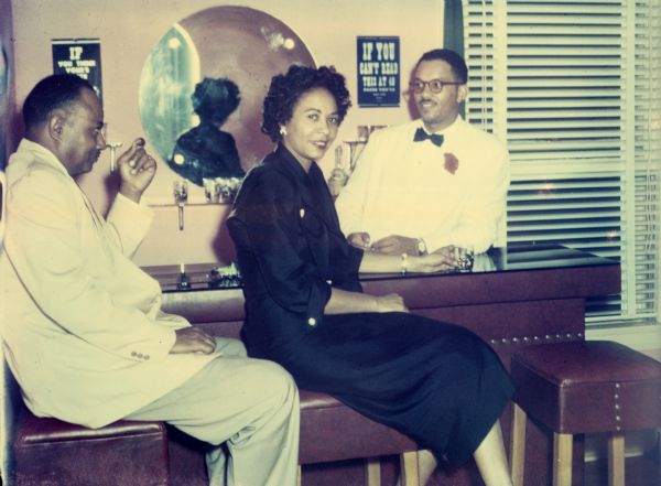 Daisy Bates is seated at a bar next to an unidentified man. She is wearing a black dress and is holding a cocktail.