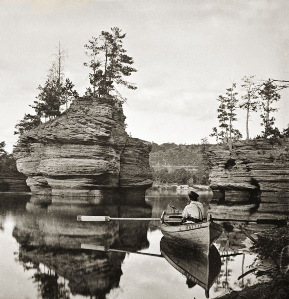 Sugar Bowl rock formation. In the foreground is a man in rowboat with the name "Hattie" painted on its side. The river is smooth as glass, and on the opposite shore is a fence on the side of a steep hill.