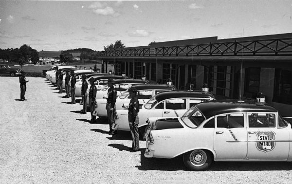 State patrol officers line up beside their patrol cars at Old District 1 State Patrol Field Headquarters on Highway 12. They appear to be receiving instructions from a superior officer. In the background is a large barn with a sign painted on its side.