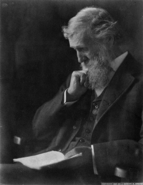 Portrait of John Muir. He is seated and is reading a book.