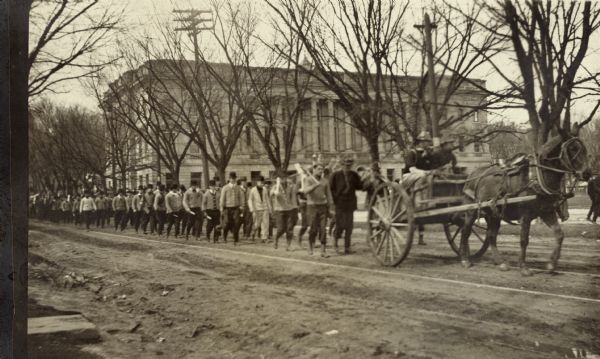 A long column of men following a horse-drawn cart on State Street. The Wisconsin Historical Society, then known as the State Historical Society of Wisconsin, is visible in the background. The man in the cart may be wearing a military uniform. Many of the men following appear to be wearing sweaters and hats, and are carrying white objects in their hands.