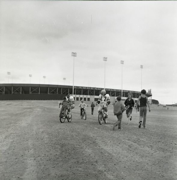 Green Bay Packers quarterbacks, Jerry Tagge (#17) and Scott Hunter (#16), ride bicycles loaned to them by young fans from Lambeau Field to their practice field during training camp. One player rides with a child on the handlebars while others jog or walk along. Lambeau Field is visible in the background.