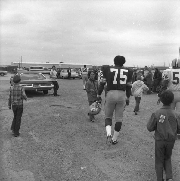 Green Bay Packers player Dave Pureifory (#75) carries his helmet as he walks among fans to the practice field. Larry McCarren (#54) can be seen ahead of him in the distance.
