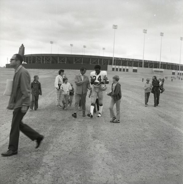Green Bay Packers running back, Ron McBride, wearing a #24 jersey and carrying a helmet, signs autographs for fans as he walks from Lambeau Field to the practice field. Lambeau Field is visible in the background.