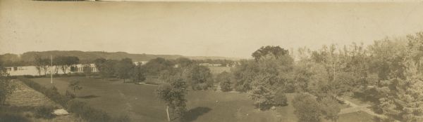 Elevated panoramic view of the west lawn at Villa Louis. Several trees and shrubs, a flag pole, and a swimming hole can be seen in the yard. A pontoon railroad bridge and the Mississippi River are visible in the distance.