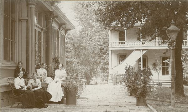 View of the east terrace at Villa Louis. The Dousman's office building is in the background. Relaxing near the home are Louis DeVierville Dousman, Judith Dousman, Virginia Dousman, Violet Dousman and an unidentified woman. Two dogs accompany the group. There is a lamppost at right near a path.