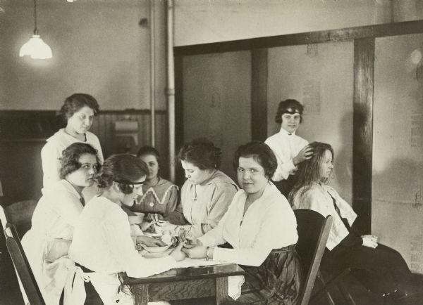 Women seated at a table learning to give manicures as part of a manicuring class. Behind the group another woman is sitting in a chair  receiving a scalp massage.