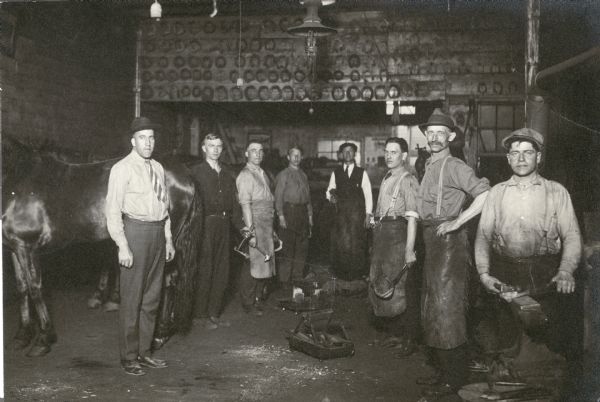 Men conscripted to learn horseshoeing pose together at the American Express Company. A horse is partially visible on the left side of the image. Rows of horseshoes are attached to a wall above the men in the background.