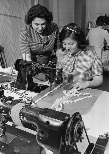 Instructor Ida Mussa (left) assists student Victoria Vega in a power sewing class at Milwaukee Vocational School. Vega is sewing an apron on a Singer sewing machine.