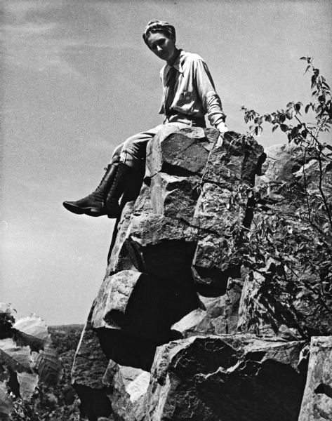 Virginia Mueller is seated on a rock formation. She is wearing tall, black boots.