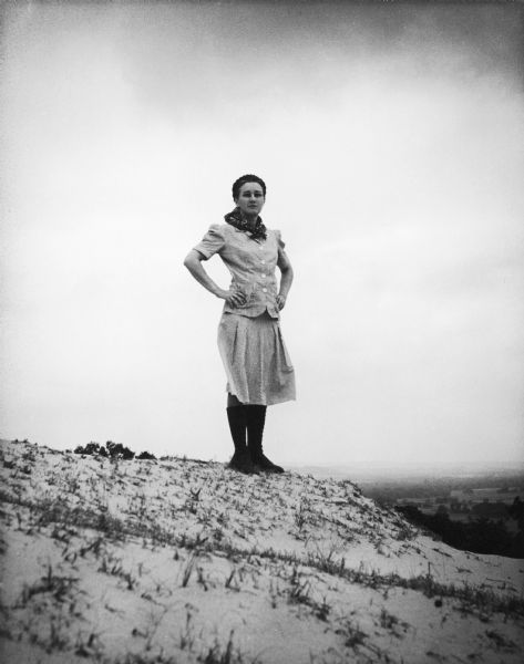 Virginia Mueller stands on a dune with her hands on her hips. In the far background is a valley with trees and fields.