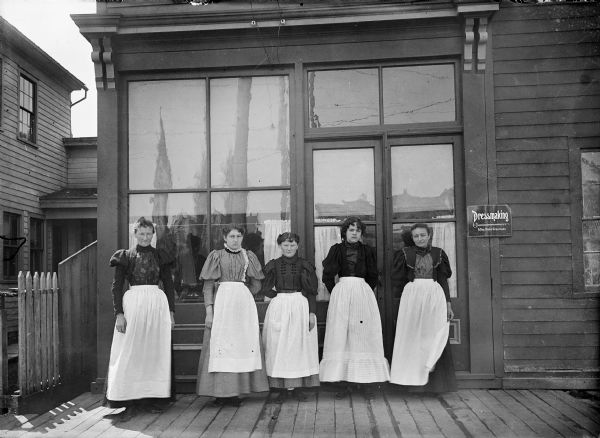 Miss Graumann, far left, poses with four dressmakers in front of her shop.  A sign on the right side of the door states "Dressmaking, Miss Annie Graumann".  She conducted the business at North Ninth Street and Michigan Avenue for five years.  <i>Pott's Directory of the City and County of Sheboygan, 1897-'98</i> lists 831 Michigan Avenue as the address of both her shop and residence.  The directory lists a total of 35 dressmakers' establishments in the city, at the time.