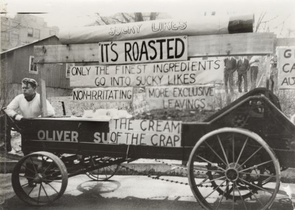 Man standing at the back of a manure spreader decorated with signs reading: "Sucky Likes," "It's Roasted," "Only the Finest Ingredients Go into Sucky Likes," "Non-Irritating," "More Exclusive Leavings," and "The Cream of the Crap."