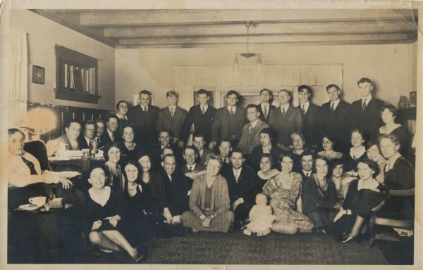 Group portrait of John R. Commons and his students during a Friday Nighters gathering at Ho Cheera. There is a doll on the floor at the front of the group near center.