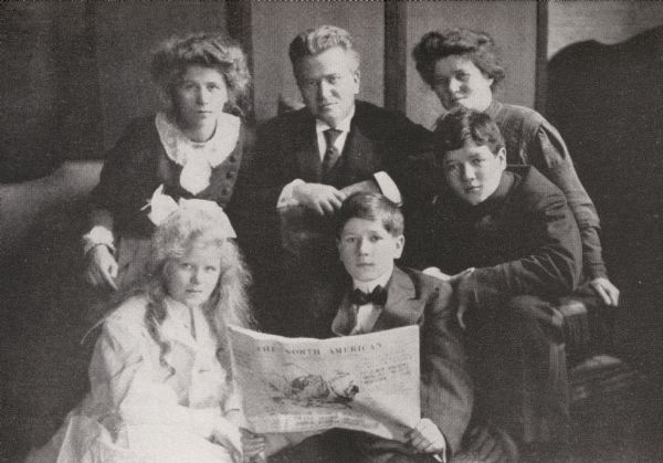 Studio portrait of the La Follette family. Clockwise from top left: Fola, Bob, Belle, Robert Jr., Philip, and Mary. Phil holds an issue of "The North American".