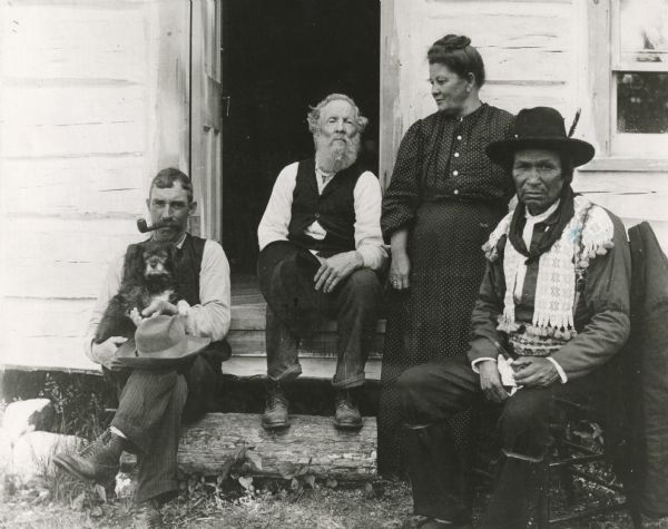 Ira Robinson Isham family at the Lac Courte Oreilles Indian Reservation. From left to right: Isham's son-in-law, Isham, his daughter, and Indian medicine man Gosh-gi-bosh. The man at left holds a dog in his lap.