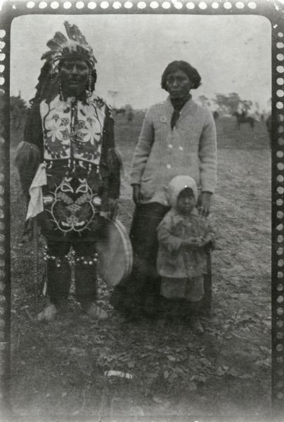 Informal portrait of Mrs. Charles Belille, standing with a man and a child (possibly her husband and their child). The man is wearing traditional Ojibwe ceremonial clothing and is holding a drum. In the field in the background are horses.