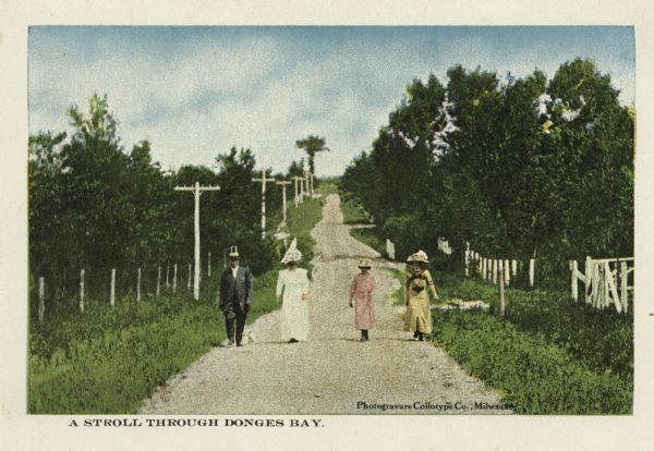 Well-dressed group of three women and one man walking along a dirt road lined with fences and telephone poles. Caption reads: "A Stroll through Donges Bay."