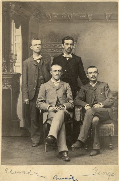 The four Burdick family brothers pose together for a studio portrait in front of a painted backdrop. Standing on the far left is Edmond. Seated at the far right is George. Though not identified on the photo the other two brothers are names Robert and Julius.