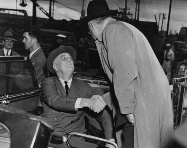 Governor Julius Heil with his back to the camera shakes the hand of President Franklin Roosevelt who was on a surprise visit to Milwaukee for a tour of the Allis-Chalmers plant.