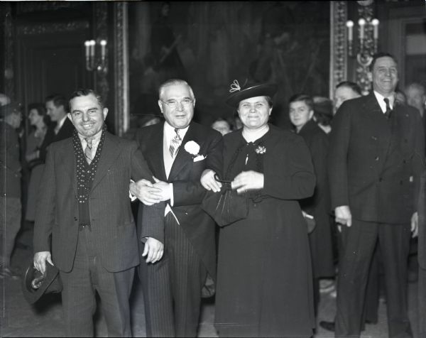 Governor Julius P. Heil, his wife and an unidentified man pose arm-in-arm in the Governor's Conference Room at the Wisconsin State Capitol. A number of others mingle in the background. A large painting is visible behind the group.
