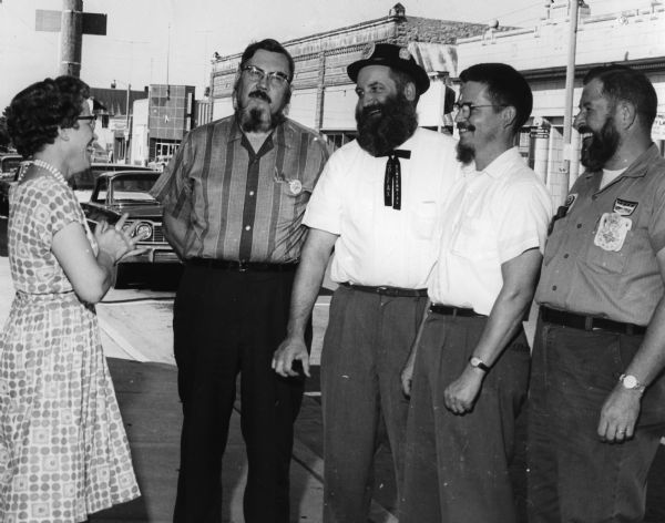 Mrs. Harold Olson examines the beards of Leon Dunbar, Louis Christianson, Dick Toycen and Perley Entzminger. They are standing on the 500 block of Main Street with the bank in the background at right. The building at center is currently Commercial Testing Labs, inc.