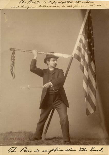 Full-length studio photograph of Louis Brosemer wearing a suit and hat. He is standing before a United States flag holding an oversized pen as though it was a spear in his right hand, and holding a sword by the blade in his left hand.