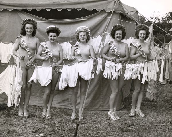 Five women circus performers in full costume pose behind a clothesline upon which bras, underwear, gloves and socks hang to dry.