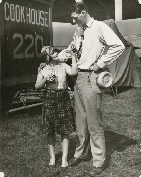 A young woman and a very tall man stand together in front of the cookhouse wagon. She is holding a bottle of soda and offering him a hotdog; he has his arm around her. They are circus employees.
