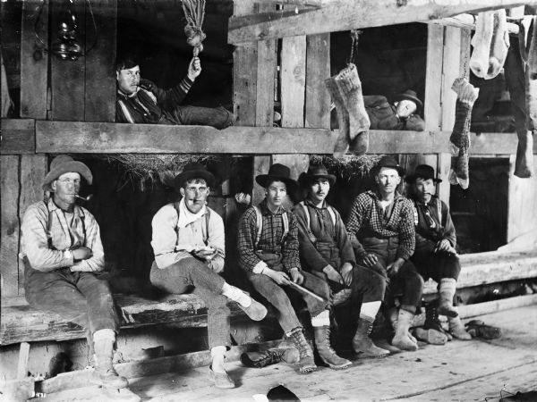 Lumbermen in their bunk house at Ole Emerson's lumber camp. Several of them have pipes in their mouths and their socks are hanging to dry.