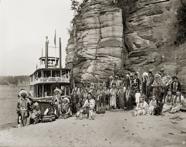 Ho-Chunk ceremonial performers posing on the sandy riverbank in front of a steamboat and rock formation.