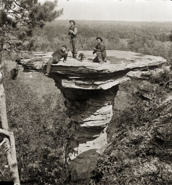 View of Stand Rock with three hunters and a dog posing on top. The man at left is sitting on the edge, the man in the middle is standing and holding a gun, and the man at right is crouching and holding a gun.