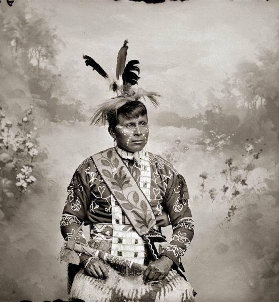 Waist-up studio portrait in front of a painted backdrop of a Ho-Chunk man, Chach-scheb-nee-nick-ah (Young Eagle) looking off to the right.