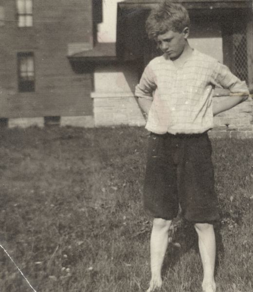 A boy in a white shirt and dark shorts stands outdoors at Hillside Home School. He has his hands on his hips and is barefoot.