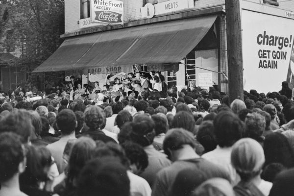A man is speaking through a megaphone to a large crowd from the steps of the Mifflin Street Co-op, 32 North Bassett Street. Possibly Attorney Melvin Greenberg, introducing Madison Mayor William Dyke. The Mayor agreed to meet with University of Wisconsin students after several days of rioting followed by police breaking up an unsanctioned student block party. A Coca-Cola sign on the front of the building still advertises its former identity as a White Front grocery store.