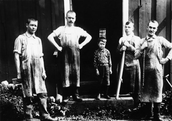 Four men in aprons and a boy stand outside a machine shop.