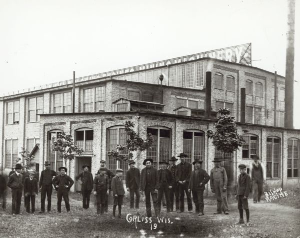 Men and boys pose outside a machine factory in Corliss. Almost all of them are similarly dressed in dark hats, jackets, and pants. A large sign is on the roof.