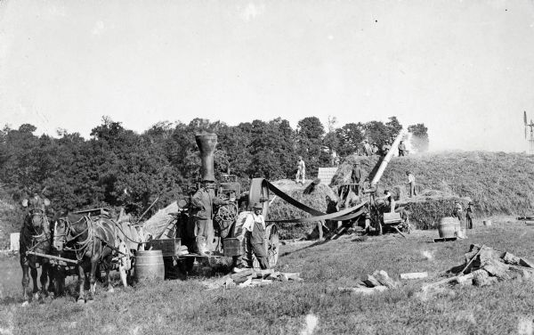 Group of men and boys threshing. They are using a tractor for a belt-driven threshing machine, and on the left two horses pull a wagon with large barrels.