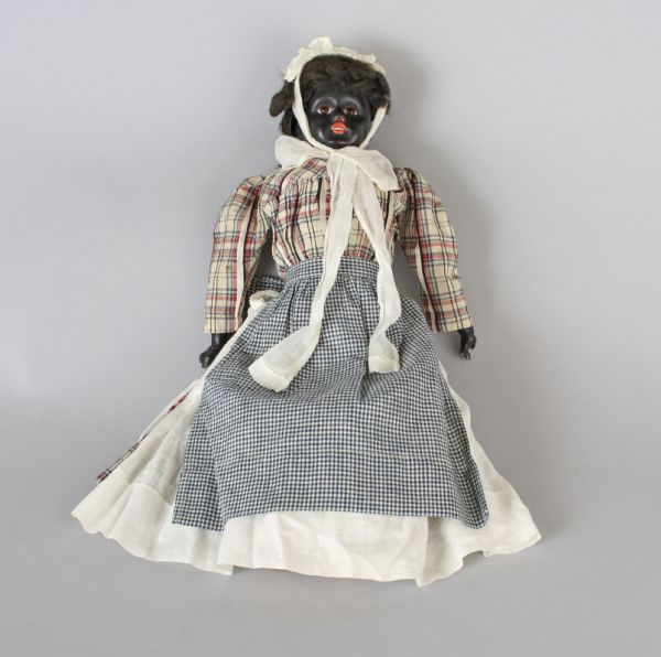 A bisque doll of a woman with her face and hands painted black. She is wearing a red plaid dress with two aprons. The doll was made in Germany in 1890-1910.