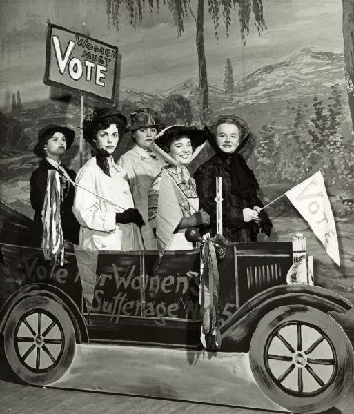 Five Civics Club members pose in a cardboard automobile in front of a painted backdrop as suffragists. They're wearing period costumes and holding "Vote" signs.