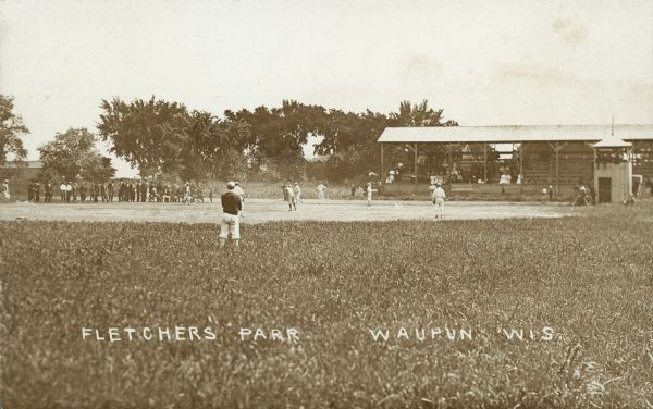 View from center field of baseball game being played in Fletcher's Park. A group of spectators are sitting under a covered grandstand on the right. Caption reads: "Fletcher's Park, Waupun, Wis."