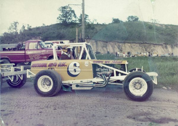 Race car number 6 sponsored by National Auto Elec. The body of the car was built by the Ideal Body Company. A man wearing a helmet and goggles is sitting in the race car. In the background are a group of men with cars parked near an eroded hill.