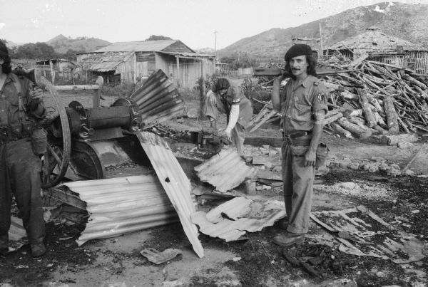 Soldiers of Fidel Castro's 26th of July Movement during the Cuban Revolution stand next to a pile of wreckage in a rural area holding guns.