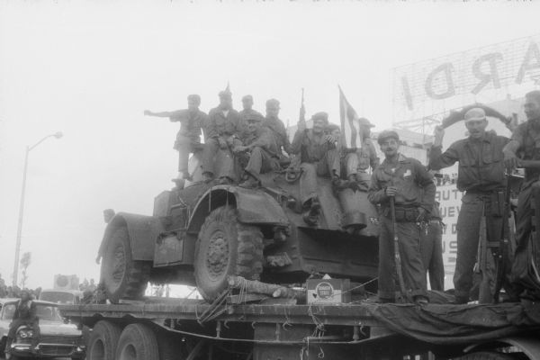 Parade celebrating Fidel Casto's arrival in Havana at the end of the Cuban Revolution. 26th of July Movement soldiers are riding on top of a military vehicle wedged in place on a trailer. Crowds and other vehicles in the parade are on the left, and large advertising signs are in the background.