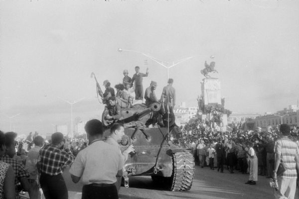 Parade celebrating Fidel Casto's arrival in Havana at the end of the Cuban Revolution.  26th of July Movement soldiers are riding on top of a captured Sherman tank as it is moving down a wide city street.  Large crowds are watching the parade from both sides of the street, including on top of an equestrian statue. Downtown city buildings are in the background.
