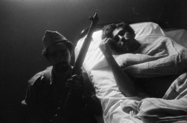 Cuban Revolution infirmary.  A soldier holding a rifle sits on the floor next to a man in bed.