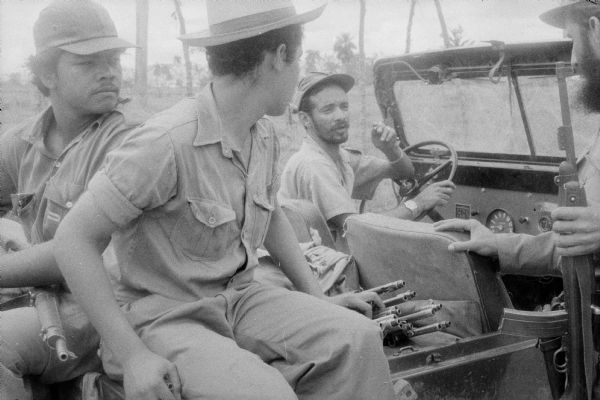 26th of July Movement soldiers during the Cuban Revolution.  Three men are around a jeep listening to the driver speak. There is a stack of firearms in the back of the jeep. Fidel Castro on right.