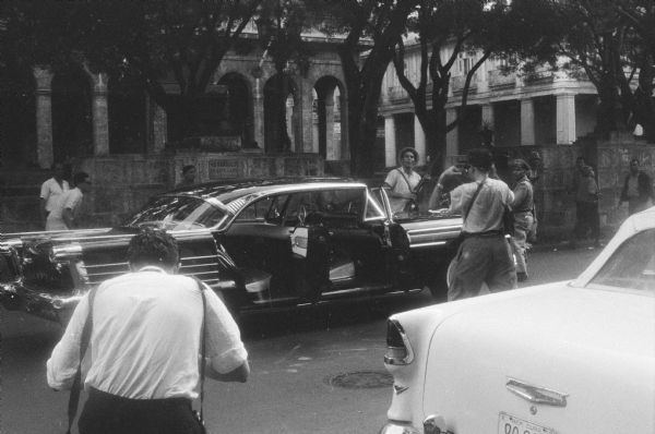 Parked limousine with open doors on a Havana street at the end of the Cuban Revolution. Two men in the foreground appear to be taking photographs of the vehicle, and men are on the sidewalk in the background. One of the men is holding a rifle.  Colonial stone buildings with arches and columns are in the background.

Possibly the limousine President Urrutia and Interior Minister Orlando rode in during the parade celebrating Fidel Castro's arrival in the city.  Castro opted to walk and ride in a jeep.