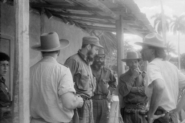 Five men, including Fidel Castro, and Celia Sanchez stand together in discussion on a shaded porch in the country during the Cuban Revolution.